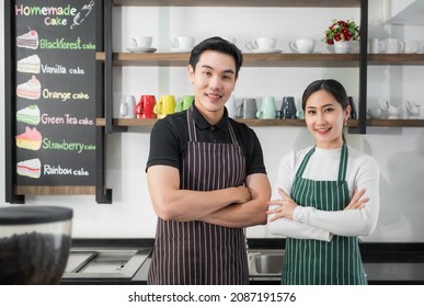 Portrait of Asian couple barista wears apron standing with arms crossed and looking at camera on counter rear in coffee shop. Young man and woman cafe owner smiling and waiting for welcome customer.