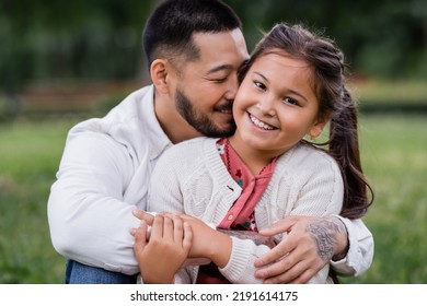 Portrait Of Asian Dad Hugging Daughter Looking At Camera In Park