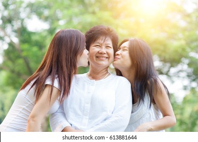 Portrait Of Asian Daughters Kissing Elderly Mother, Senior Adult Woman And Grown Child. Outdoors Family At Nature Park With Beautiful Sun Flare.