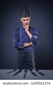 A Portrait Of An Asian Executive Chef In His Blue Uniform, Black Apron And Black Hat On Dark Grey Background With His Knives