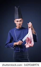 A Portrait Of An Asian Executive Chef In His Blue Uniform, Black Apron And Black Hat On Grey Background Holding A Lamb Leg