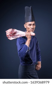 A Portrait Of An Asian Executive Chef In His Blue Uniform, Black Apron And Black Hat On Grey Background Holding A Lamb Leg