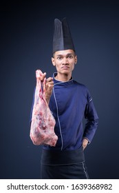 A Portrait Of An Asian Executive Chef In His Blue Uniform, Black Apron And Black Hat On Grey Background Holding A Lamb Leg