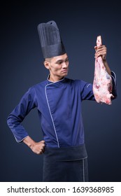 A Portrait Of An Asian Executive Chef In His Blue Uniform, Black Apron And Black Hat On Grey Background Holding A Lamb Leg