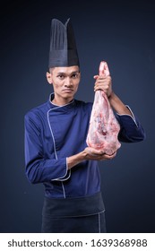 A Portrait Of An Asian Executive Chef In His Blue Uniform, Black Apron And Black Hat On Grey Background Holding A Lamb Leg