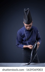 A Portrait Of An Asian Executive Chef In His Blue Uniform, Black Apron And Black Hat On Grey Background With A Skipjack Tuna