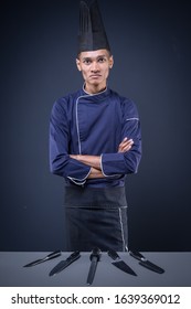 A Portrait Of An Asian Executive Chef In His Blue Uniform, Black Apron And Black Hat On Dark Grey Background With His Knives