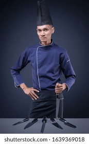 A Portrait Of An Asian Executive Chef In His Blue Uniform, Black Apron And Black Hat On Dark Grey Background With His Knives