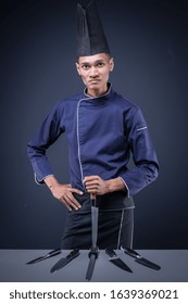 A Portrait Of An Asian Executive Chef In His Blue Uniform, Black Apron And Black Hat On Dark Grey Background With His Knives