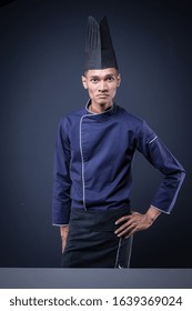 A Portrait Of An Asian Executive Chef In His Blue Uniform, Black Apron And Black Hat On Dark Grey Background