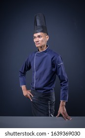 A Portrait Of An Asian Executive Chef In His Blue Uniform, Black Apron And Black Hat On Dark Grey Background