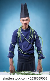 A Portrait Of An Asian Executive Chef In His Blue Uniform, Black Apron And Black Hat On Blue Background With Long Beans On Him