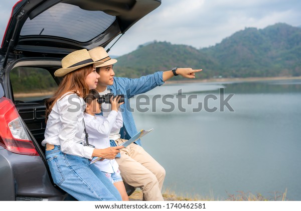 Portrait of Asian family sitting in car with\
father pointing to view and mother holding maps with daughter\
looking beautiful landscape through binoculars while vacation\
together in holiday.