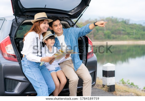 Portrait of Asian family sitting in car with father\
pointing to view and mother with daughter looking beautiful\
landscape and holding maps while vacation together in holiday.\
Happy family time.
