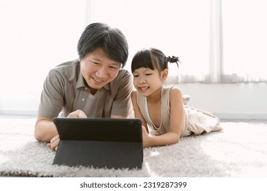 Portrait of Asian father and cute little daughter watching cartoons using digital tablet gadget e-books for kids on-line lying on carpet at home. Parental control protect from bad content concept - Powered by Shutterstock