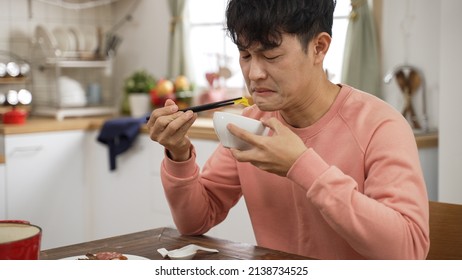 Portrait Of Asian Father Making Expression Of Disgust While Tasting Food With Chopsticks And Bowl At Home Dining Table Alone.