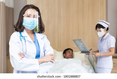 Portrait Of Asian Female Doctor Wearing Medical Protective Face Shield And Mask Looking At Camera In Arm Crossed. Nurse Taking Care Of Patient Sickness On Bed In Hospital Ward. New Normal Concept