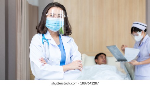 Portrait Of Asian Female Doctor Wearing Medical Protective Face Shield And Mask Looking At Camera In Arm Crossed. Nurse Taking Care Of Patient Sickness On Bed In Hospital Ward. New Normal Concept