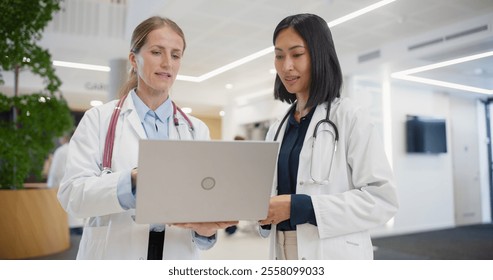 Portrait of an Asian Female Physician Discussing a Medical Case with a Middle Aged Doctor, Referencing Notes on a Laptop Computer while Standing Together in a Busy Hospital Lobby - Powered by Shutterstock