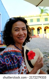 Portrait Of An Asian Female Tourist In Floral Dress, Holding A Panama Hat, Giving A Big Smile At The Plaza Mayor In Trinidad, Cuba.