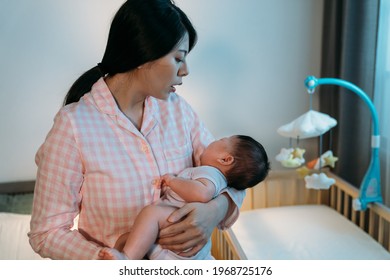 Portrait Asian First Time Mom In Pajamas Is Holding And Consoling Her Newborn Child  By The Baby Bed At Midnight In The Bedroom At Home.