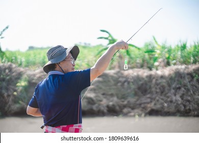 Portrait Of Asian Fisherman Catching Fish With Reel Rod And Fishing Lure Or Fake Bait At Canal .Asian Mature Man Fishing At Canal Or River.