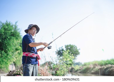 Portrait Of Asian Fisherman Catching Fish With Reel Rod And Fishing Lure Or Fake Bait At Canal .Asian Mature Man Fishing At Canal Or River.