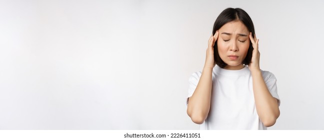 Portrait of asian girl feeling headache, migraine or being ill, standing in white t-shirt over white background. Copy space