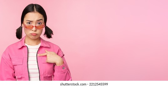 Portrait Of Asian Girl Looks Confused And Points At Herself, Perplexed Face, Stares With Disbelief At Camera, Stands Over Pink Background