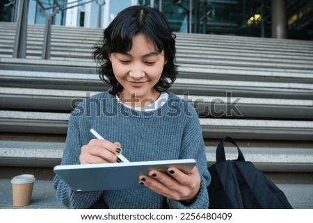 Portrait of asian girl student, hipster sitting on stairs with digital tablet and cup of coffee, draws digital art, makes design project.