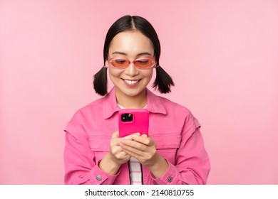 Portrait of asian girl in sunglasses using smartphone. Woman looking at mobile phone, browsing in app, standing over pink background
