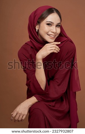 Portrait of Asian girl wearing maroon dress and turban style hijab sitting on a chair isolated over dark background. Stylish Muslim female fashion lifestyle concept.