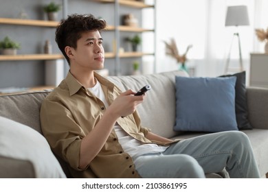 Portrait Of Asian Guy Watching TV Show Or Film, Holding Remote Control, Switching Channels. Young Man Spending Weekend Free Time Sitting On Comfortable Sofa At Home In Living Room, Copy Space