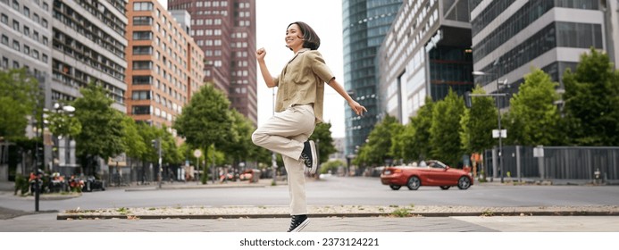 Portrait of asian happy girl jumping and dancing in city centre, posing on streets, express joy and excitement. Copy space - Powered by Shutterstock