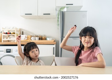 Portrait Of Asian Happy Little Cute Kid Holding Chocolate In Kitchen. Young Lovely Preschool Child Sibling Sister Sit On Table Enjoy Eating Sweet Chocolate Bar And Smiling, Looking At Camera At Home.