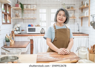 Portrait Of Asian Happy Senior Elderly Woman Standing In Kitchen At House Feeling Happy And Enjoy Retirement Life. Attractive Older Grandmother With Apron Stay At Home, Smiling And Looking At Camera.