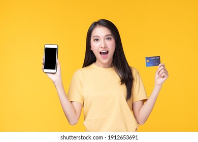 Portrait Asian Happy Young Girl Smiling Cheerful And Showing Plastic Credit Card While Holding Mobile Phone Isolated On Yellow Background