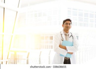 Portrait Of Asian Indian Medical Doctor Holding File Folder, Standing Outside Hospital Building Block, Beautiful Golden Sunlight At Background.