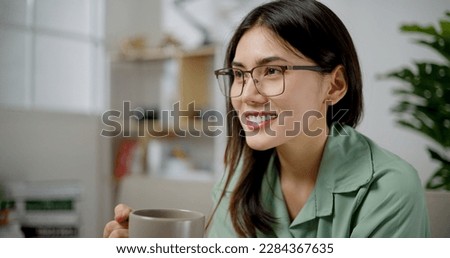 Portrait of Asian joyful young woman wearing glasses enjoying a cup of coffee at home. Smiling pretty girl drinking hot tea. People and lifestyle concepts.