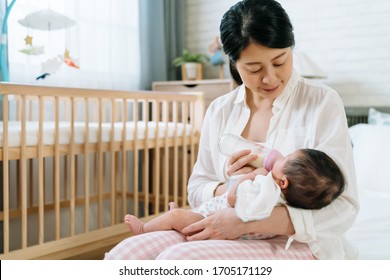 portrait asian lady lowering her head is sitting in bedroom feeding her baby. chinese mom looking her young child lovingly while giving her milk. mother's day concept.