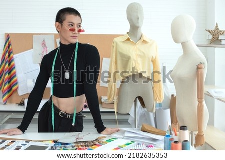 Portrait of Asian LGBTQ guy at working desk with tape measure on the neck, mannequin nearby. small business entrepreneur dressmaker and fashion designer concept
