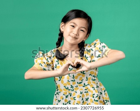 A portrait of Asian litte girl wearing a fruit-patterned dress, making a heart symbol with both hands, isolated on a green background.