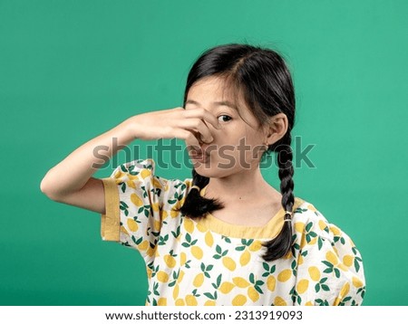 A portrait of Asian litte girl wearing a fruit-patterned dress, seen smelling something and holding her nose in disgust, isolated on a green background.