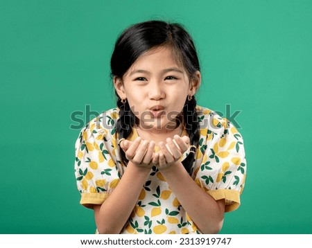 A portrait of Asian litte girl wearing a fruit-patterned dress, seen blowing on both of her hands, isolated on a green background.