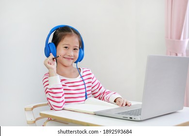 Portrait of Asian little child wearing headphone with holding pencil looking at camera study online learning class with laptop computer during new normal Covid-19 coronavirus at home