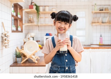 Portrait Of Asian Little Cute Kid Holding A Cup Of Milk In Kitchen In House. Young Preschool Child Girl Or Daughter Stay Home With Smiling Face, Feel Happy Enjoy Drinking Milk And Then Look At Camera.