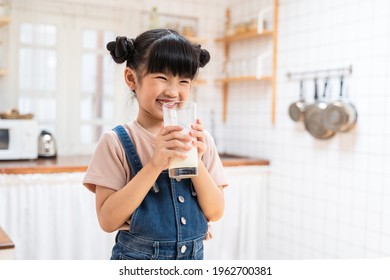 Portrait Of Asian Little Cute Kid Holding A Cup Of Milk In Kitchen In House. Young Preschool Child Girl Or Daughter Stay Home With Smiling Face, Feel Happy Enjoy Drinking Milk And Then Look At Camera.