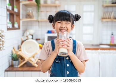 Portrait Of Asian Little Cute Kid Holding A Cup Of Milk In Kitchen In House. Young Preschool Child Girl Or Daughter Stay Home With Smiling Face, Feel Happy Enjoy Drinking Milk And Then Look At Camera.