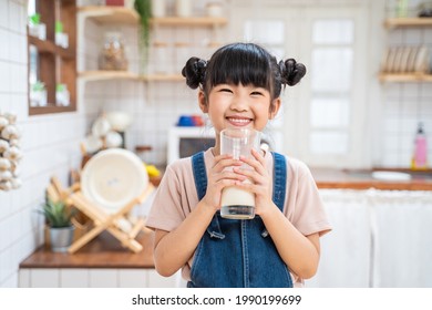 Portrait Of Asian Little Cute Kid Holding A Cup Of Milk In Kitchen In House. Young Preschool Child Girl Or Daughter Stay Home With Smiling Face, Feel Happy Enjoy Drinking Milk And Then Look At Camera.