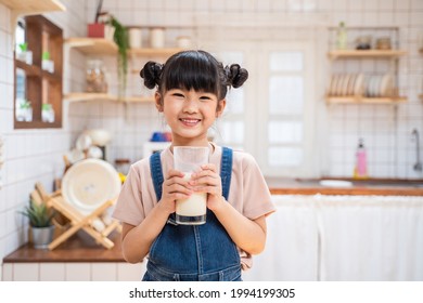 Portrait Of Asian Little Cute Kid Holding A Cup Of Milk In Kitchen In House. Young Preschool Child Girl Or Daughter Stay Home With Smiling Face, Feel Happy Enjoy Drinking Milk And Then Look At Camera.